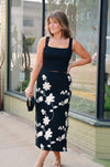 Woman wearing a black sleeveless top and floral skirt standing in front of a store window.