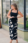 Woman wearing a black tank top and floral skirt standing outside a store.